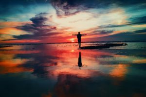 backlit person with arms outstretched against colorful sky reflected on water, photo by Mohamed Nohassi